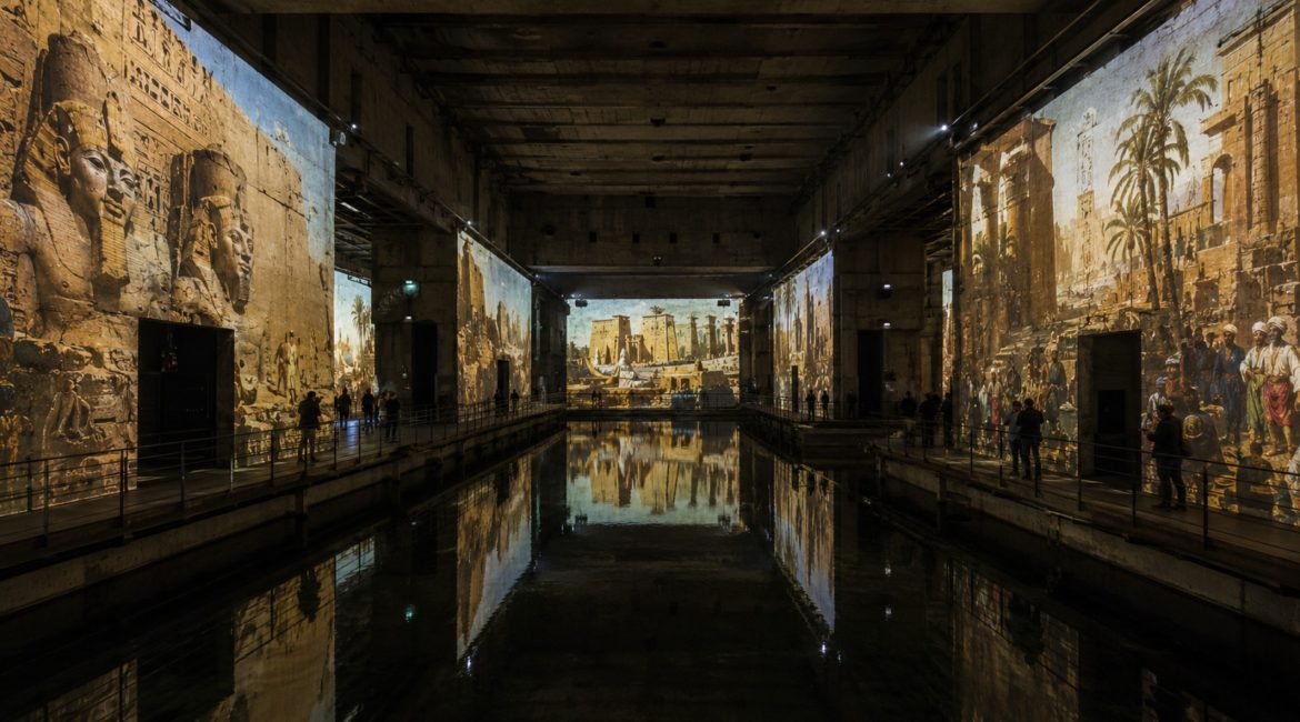 Projection monumentale de temples égyptiens et de scènes orientalistes sur les murs de béton des Bassins des Lumières à Bordeaux, reflétée dans l'eau du bassin