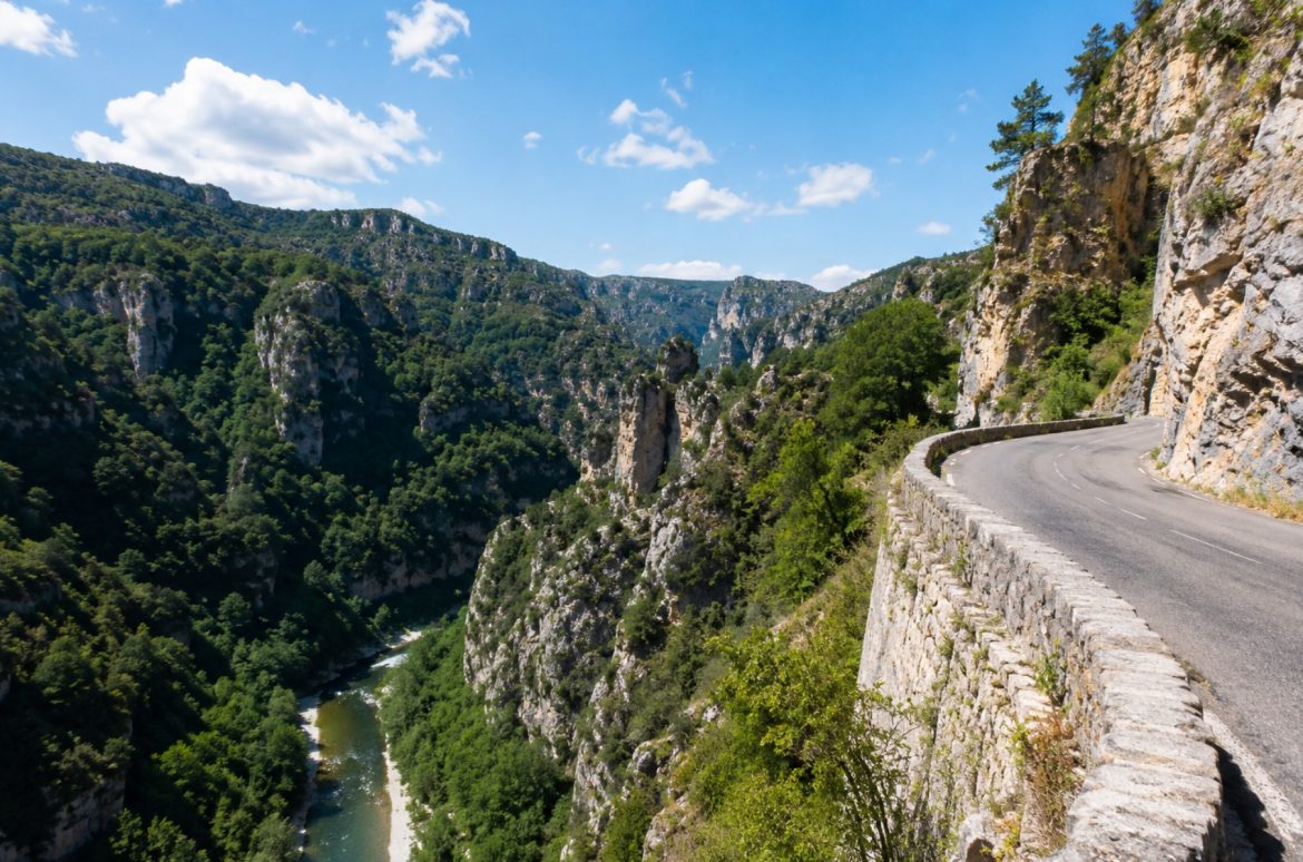 Route en corniche bordée d'un muret de pierre surplombant les Gorges du Tarn, la rivière encaissée entre les falaises calcaires