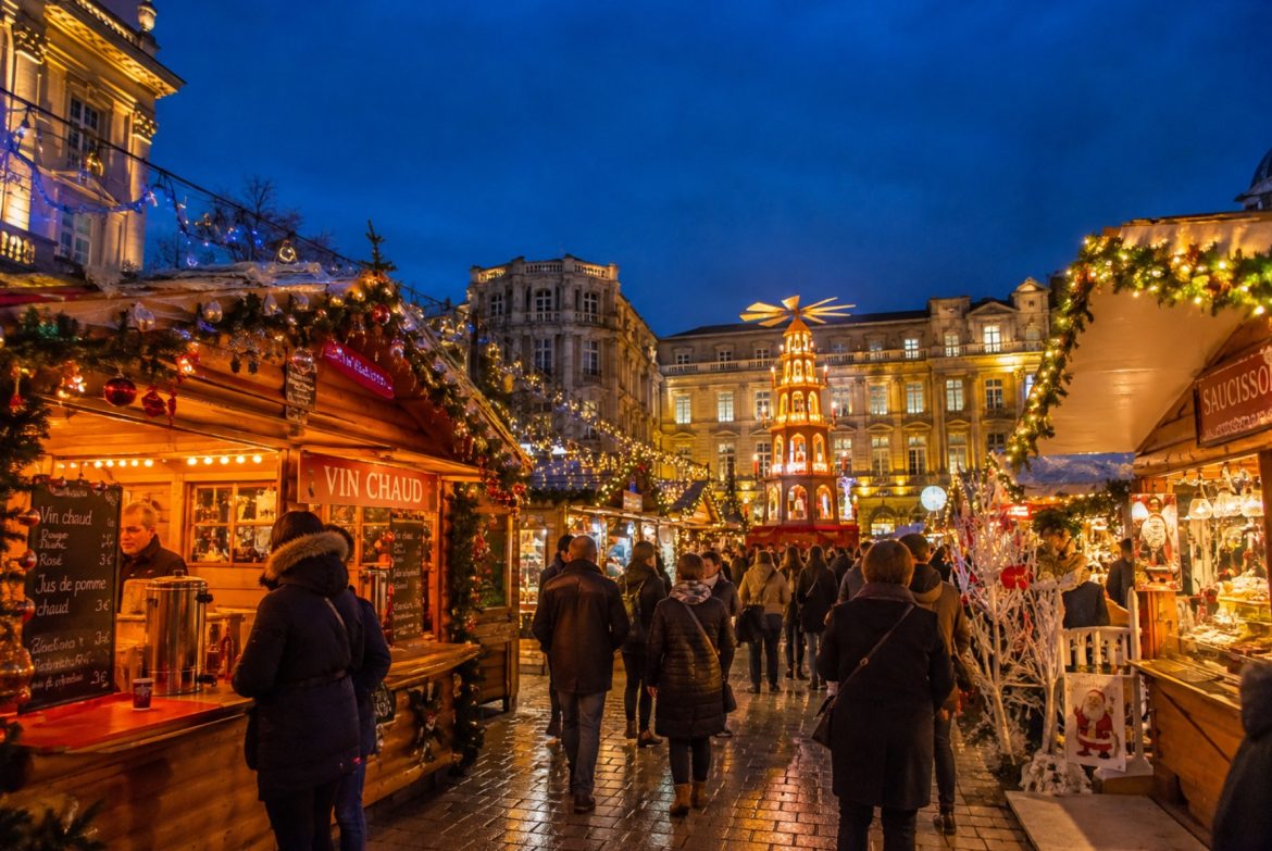Chalets en bois illuminés d'un marché de Noël à la tombée de la nuit, stand de vin chaud, guirlandes lumineuses et promeneurs emmitouflés